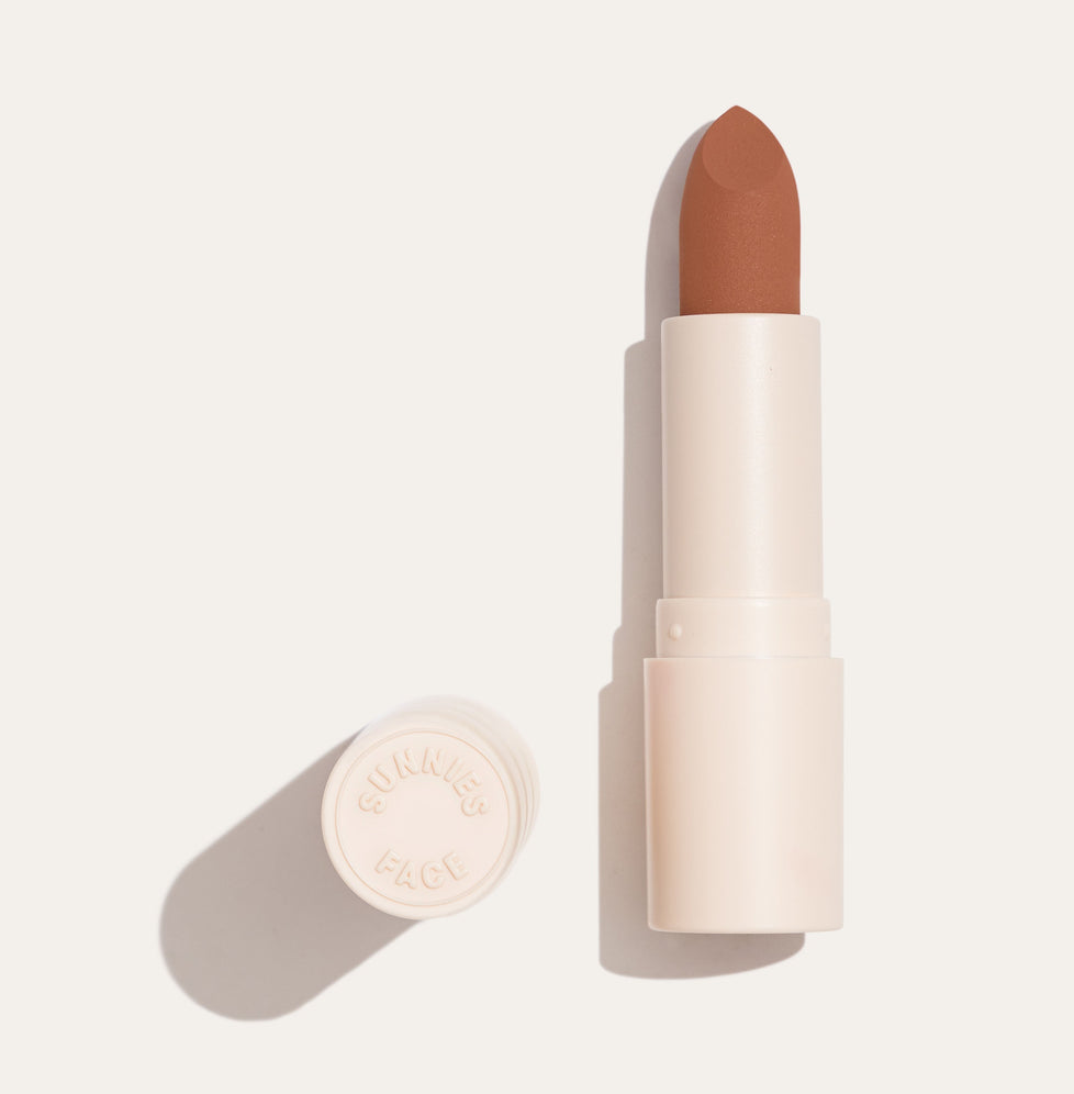 Lottiee Duty Organic Waterproof Matte Lipstick in a nude brown shade. The lipstick is shown in a sleek, cream-colored case, partially opened, revealing the smooth bullet inside.