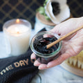 Lottiee Duty Charcoal Purifying Mask in a clear glass jar, featuring a black, creamy texture. A bamboo stick is shown mixing the mask. The jar is placed next to a lit candle and a soft towel, indicating a skincare routine.