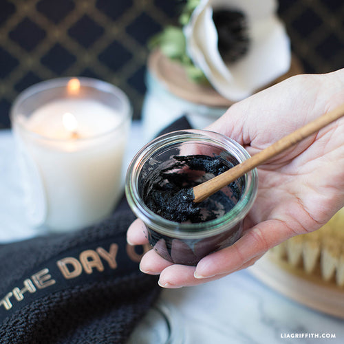 Lottiee Duty Charcoal Purifying Mask in a clear glass jar, featuring a black, creamy texture. A bamboo stick is shown mixing the mask. The jar is placed next to a lit candle and a soft towel, indicating a skincare routine.