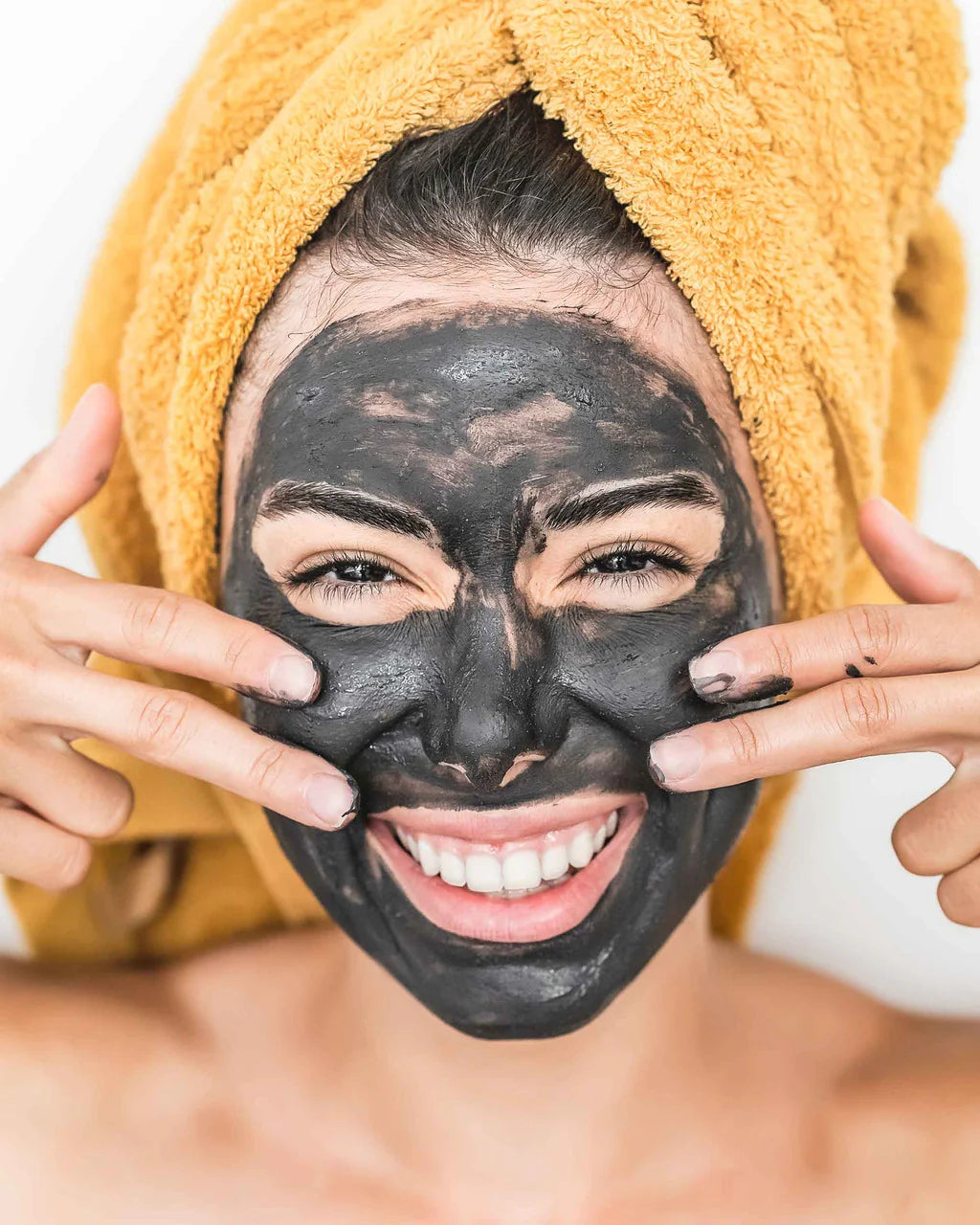 Lottiee Duty Charcoal Purifying Mask displayed on a woman’s face, showing its dark black color. The woman has a towel wrapped around her head and is smiling, indicating use of the mask for skincare.