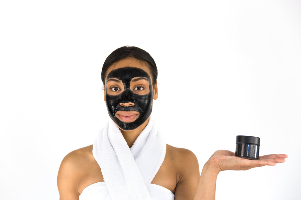 A model with a black charcoal face mask is holding a small jar labeled "Charcoal Purifying Mask" by Lottiee Duty. The model is wrapped in a white towel, highlighting the mask on her face against a clean, white background.