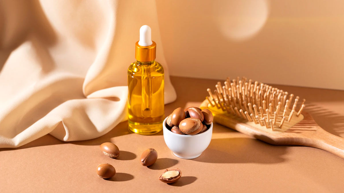Lottiee Duty Pure Argan Oil in a clear glass dropper bottle with an amber yellow hue, placed beside a small white bowl of argan nuts, a wooden hair comb, and a neutral fabric drape.