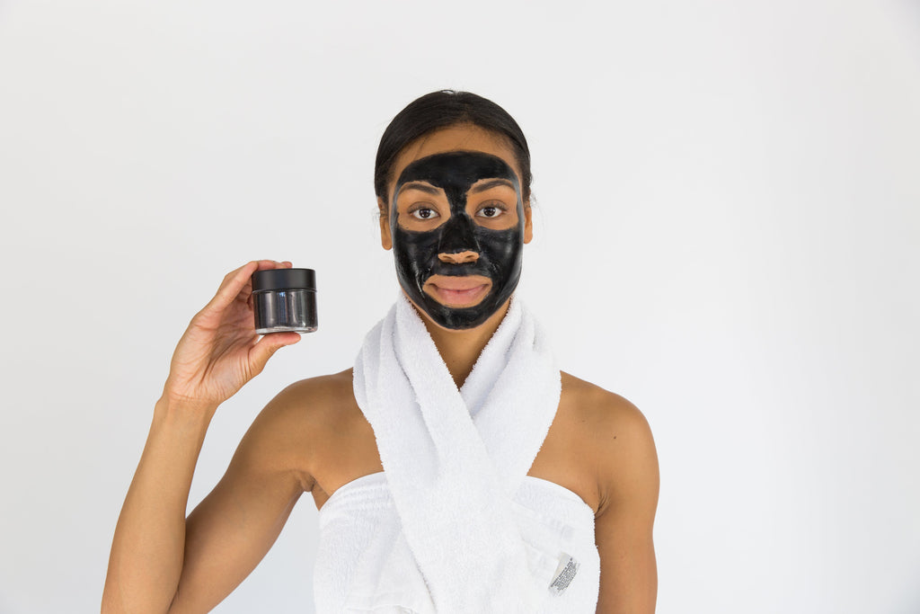 Lottiee Duty Charcoal Purifying Mask in a small black jar, held by a model with a full black facial mask. The model is wrapped in a white towel, showcasing the product for skincare use.