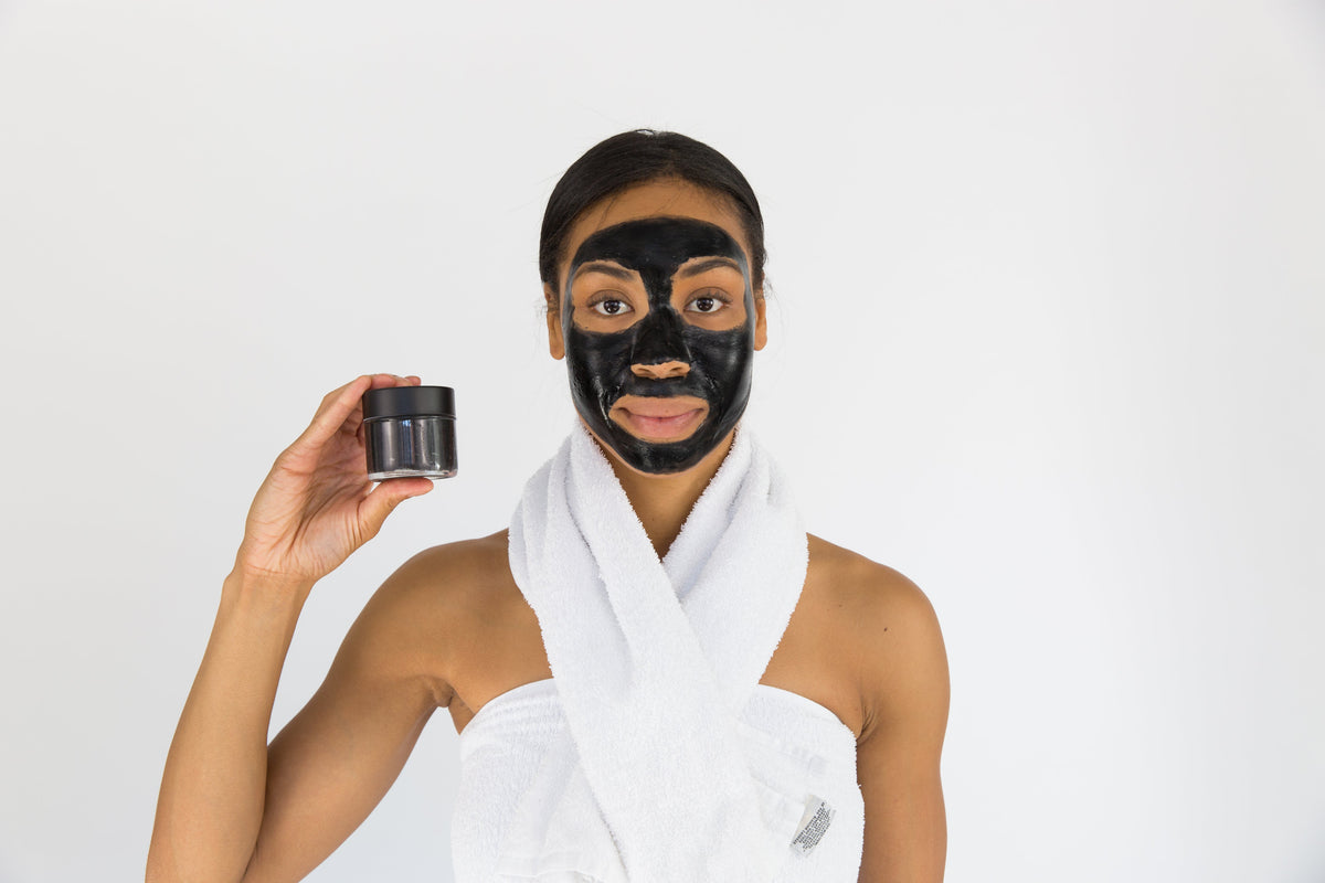 Lottiee Duty Charcoal Purifying Mask in a small black jar, held by a model with a full black facial mask. The model is wrapped in a white towel, showcasing the product for skincare use.