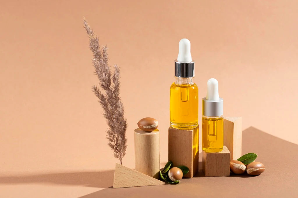 Lottiee Duty Pure Argan Oil shown in two glass dropper bottles, with the larger bottle featuring a silver cap and the smaller with a white cap. The oil is a yellow tint, displayed with wooden geometric shapes and green leaves for context.