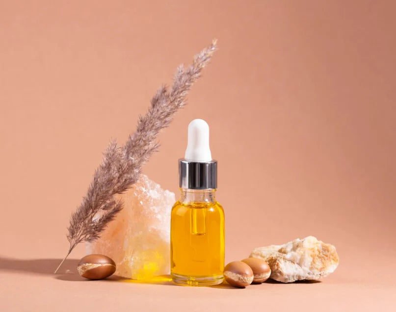 Lottiee Duty Pure Argan Oil in a glass bottle with a dropper cap, showcasing a golden yellow oil. The bottle is positioned among natural stones and dried grass on a soft beige background.