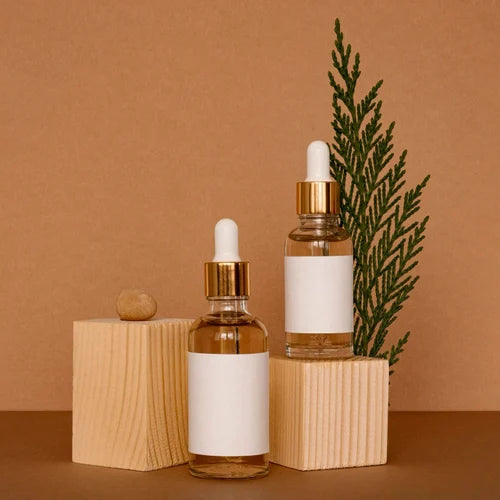 Lottiee Duty Hydrate & Luminate Elixir Serum in clear glass bottles with gold droppers, displayed on wooden blocks. The background features a brown surface and a green sprig, highlighting the product's minimalist design.